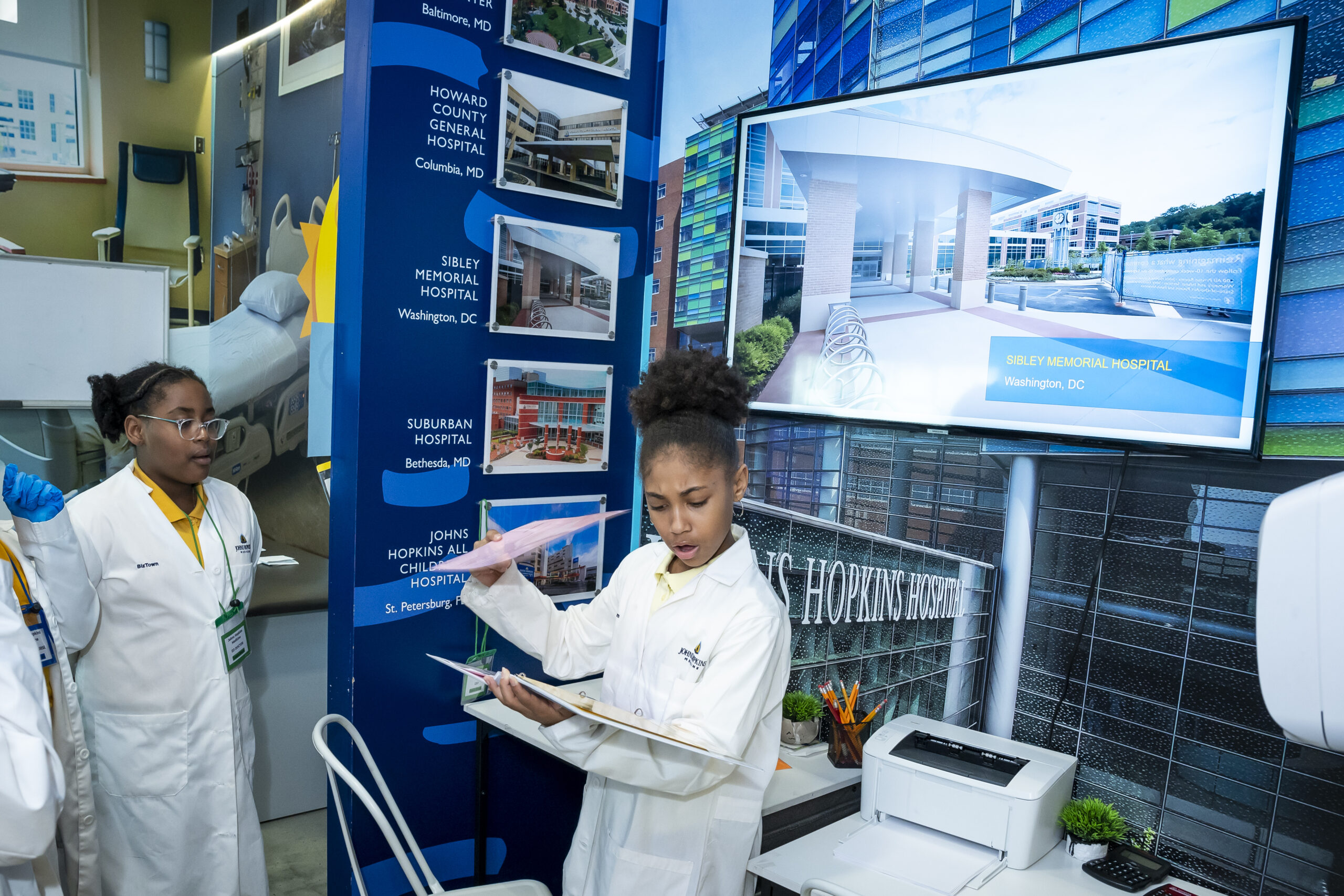Two girls in lab coats interacting with medical display and clipboard at education event