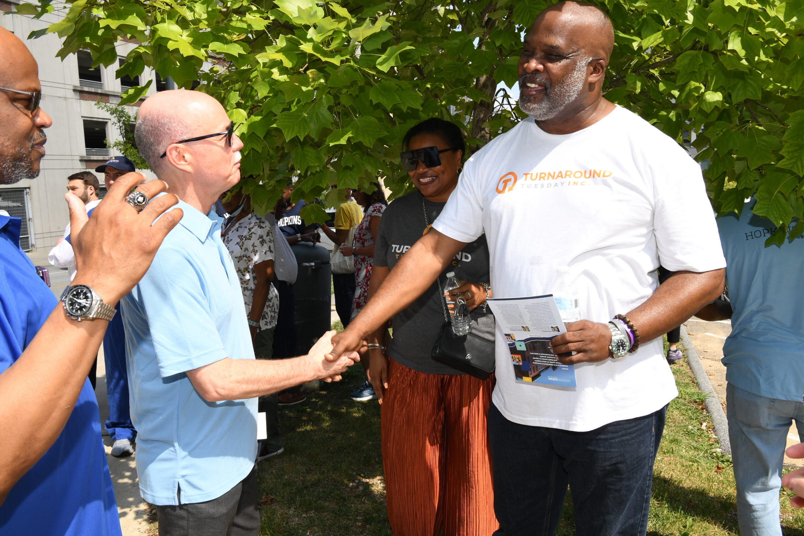 Two men shake hands under a tree, with a woman smiling nearby.