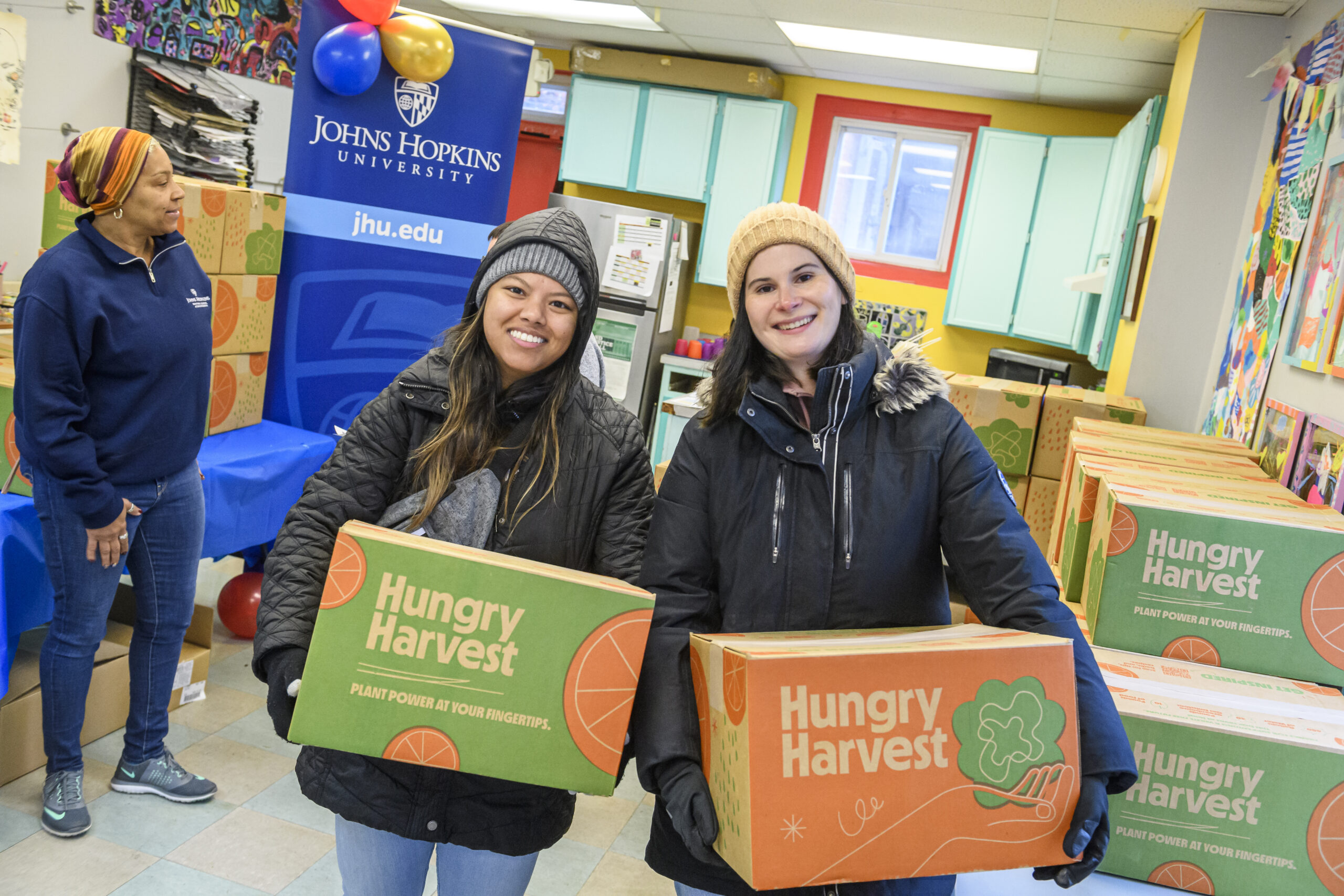 Two volunteers hold boxes labeled "Hungry Harvest" in a colorful room with a Johns Hopkins University banner.