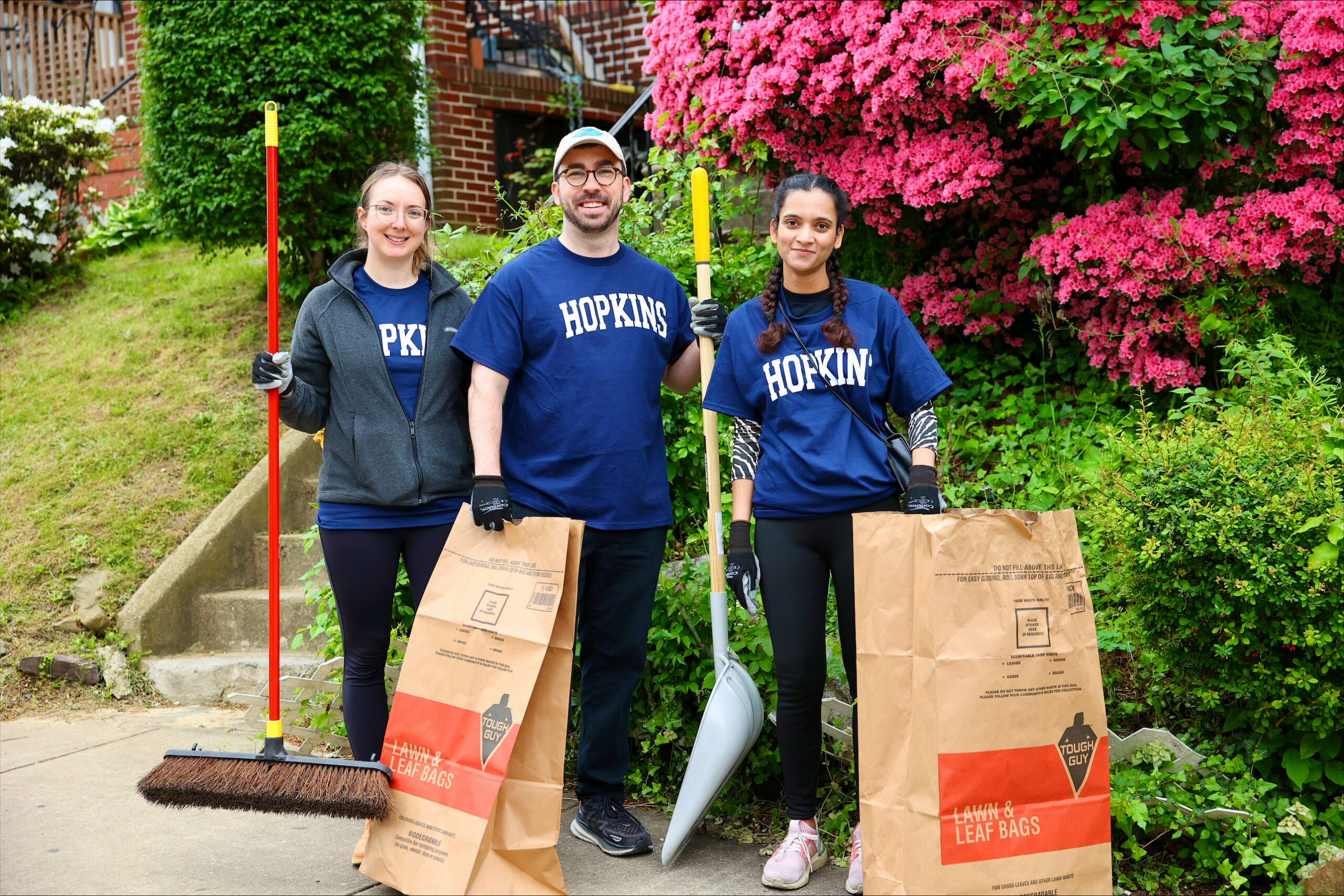 Three volunteers holding gardening tools and leaf bags, with pink flowers in the background.